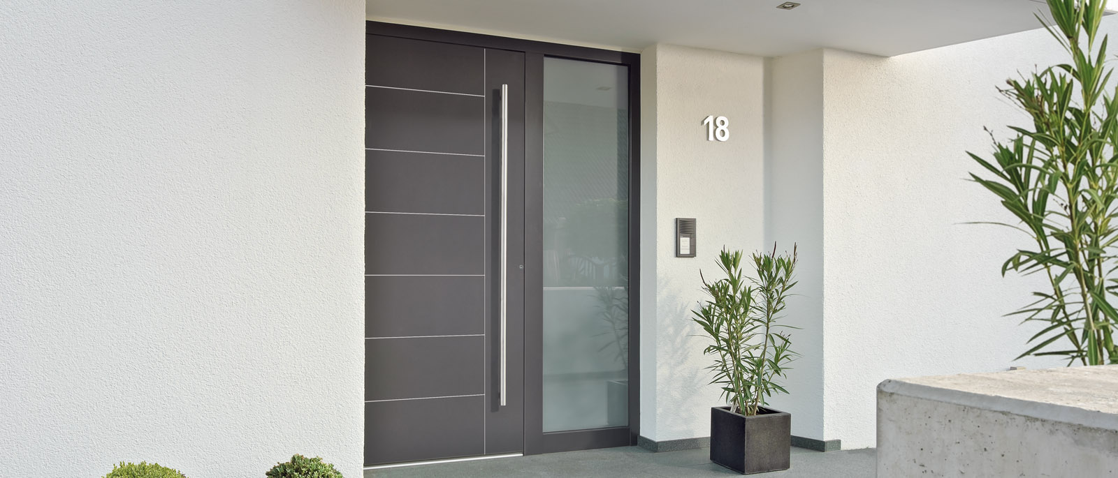 Moderne Haustür in Anthrazit mit senkrechtem Griff und Hausnummer 18 auf weißer Wand